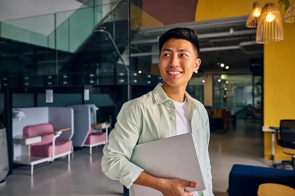 Portrait of a cheerful young asian man holding a laptop in a modern office environment
